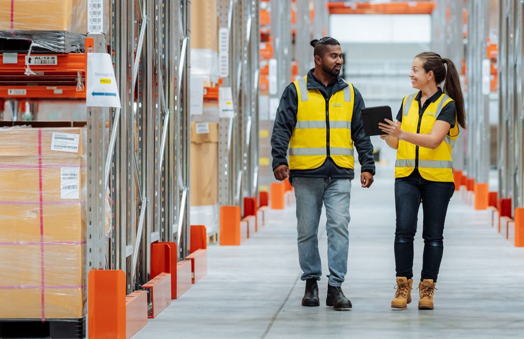 Two people walking through the warehouse wearing high-vis vests. One person is pointing to a tablet screen.
