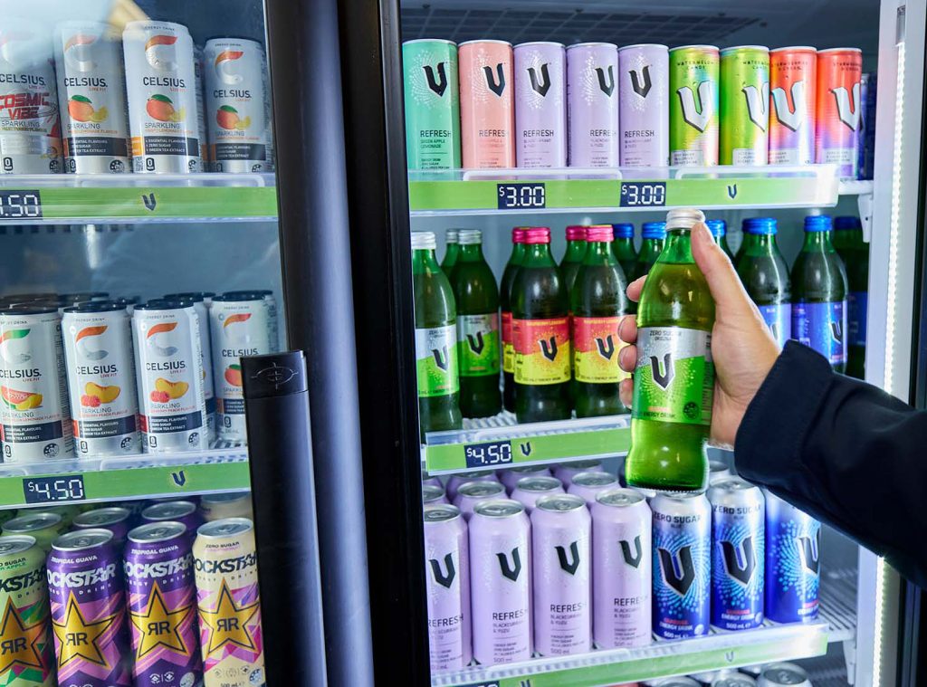 A hand holding a bottle of V Energy drink in front of a drink fridge.
