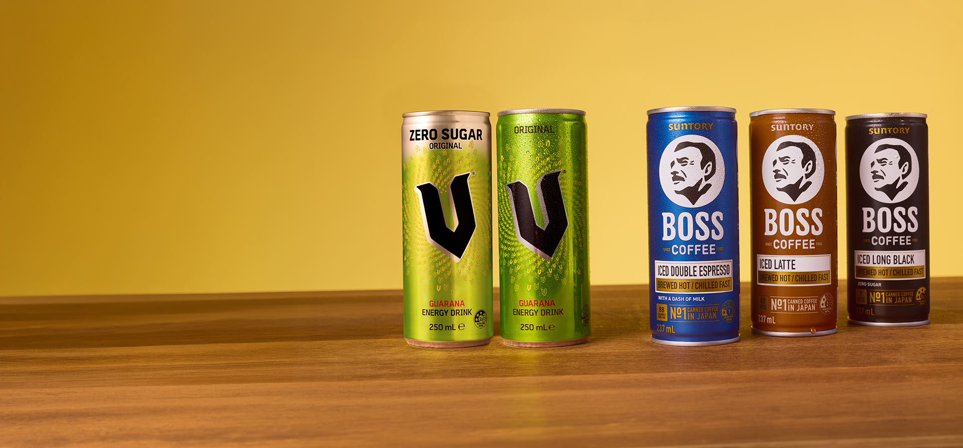 Cans of V Energy Drink and Suntory BOSS Coffee on a wooden table with a yellow wall behind.