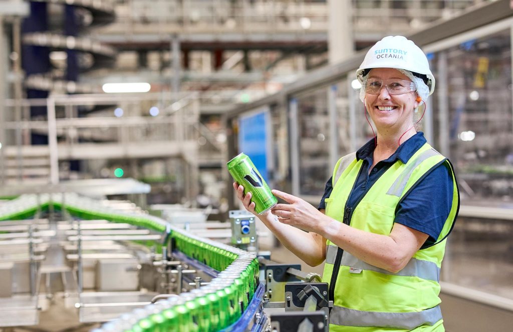 Worker in Suntory Oceania hard hat holding a can of V energy drink in front of the production line