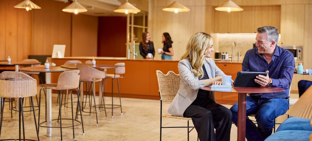A man and a woman chatting at a table in office lunch area. The man is holding a tablet.